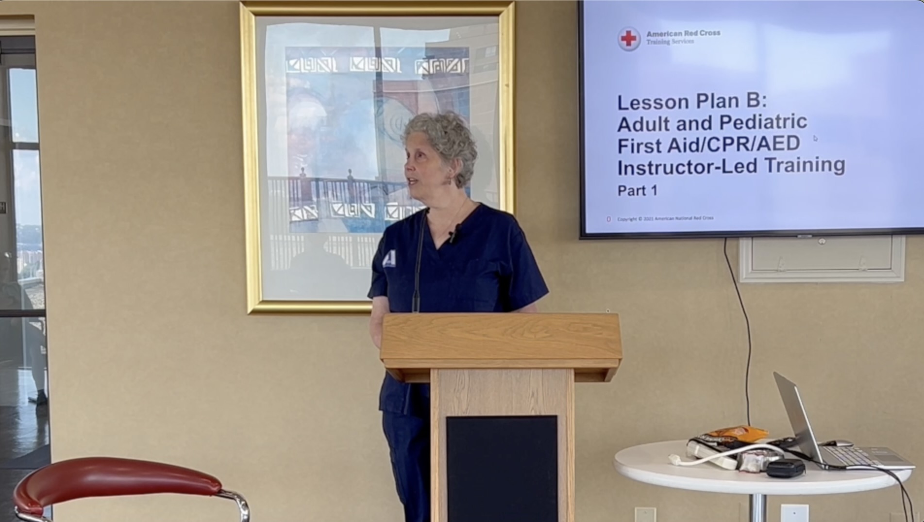 Christine, a white female with curly salt and pepper hair dressed in blue scrubs stands and a podium with a microphone. On the TV screen behind her a slide from the American Red Cross